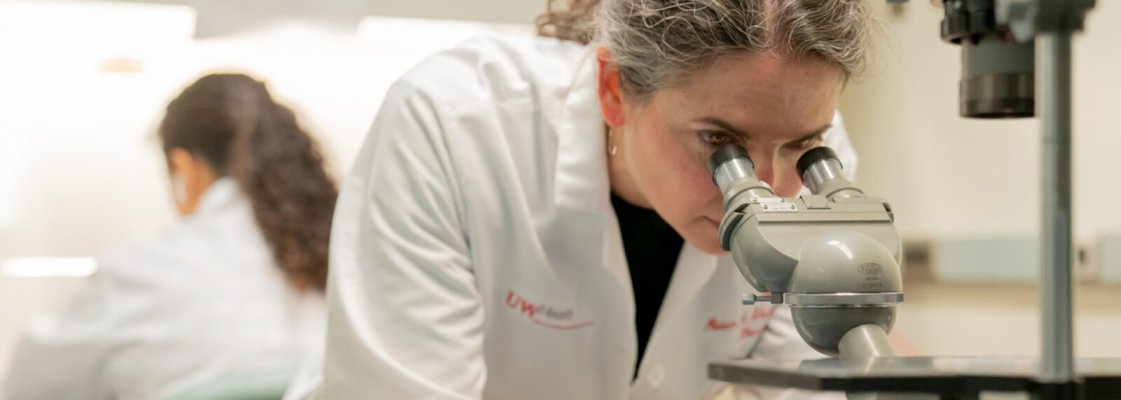 A UW Health researcher in a white lab coat looks into a microscope, with a colleague working in the background.