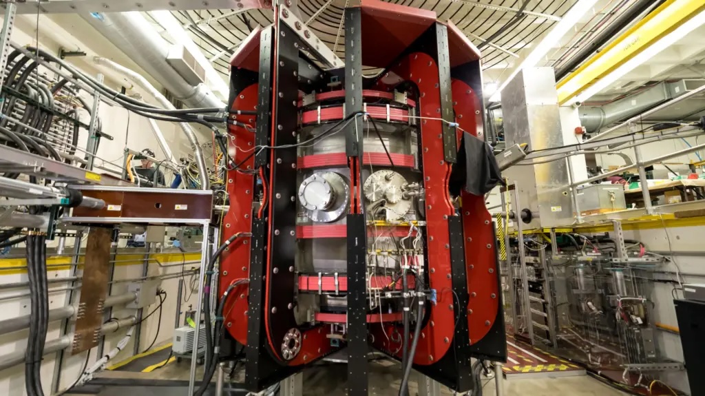 The Pegasus-III spherical tokamak, its cylindrical vacuum vessel wrapped in red magnet coils inside a black-and-red steel frame, in the UW–Madison fusion laboratory.