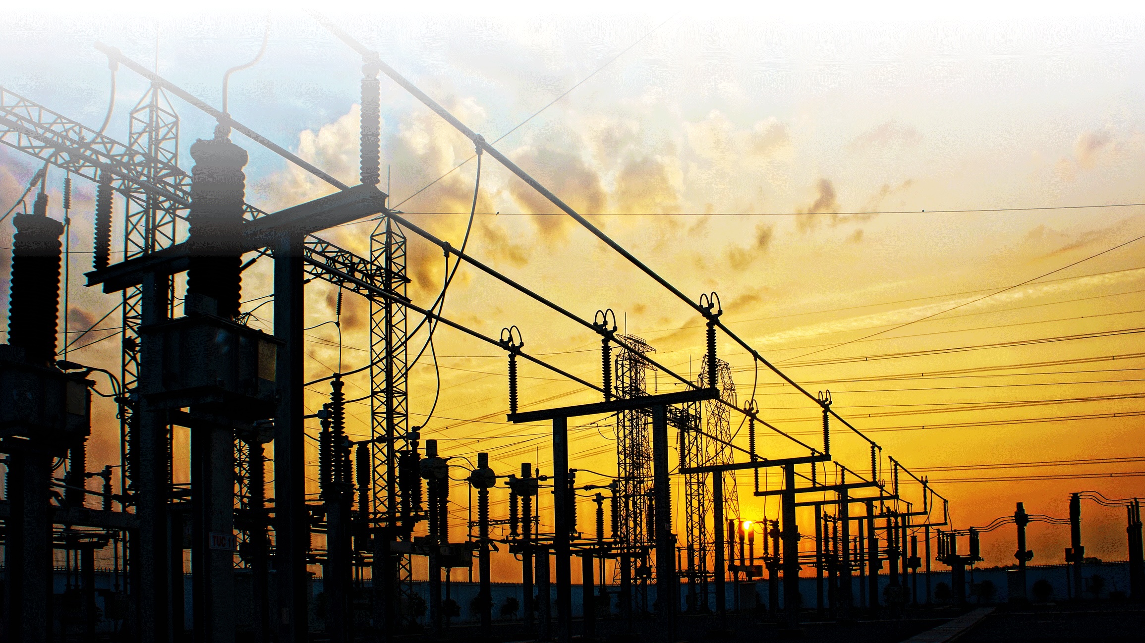 Silhouettes of high-voltage transformers, insulators, and power lines at an electrical substation against an orange sunset.