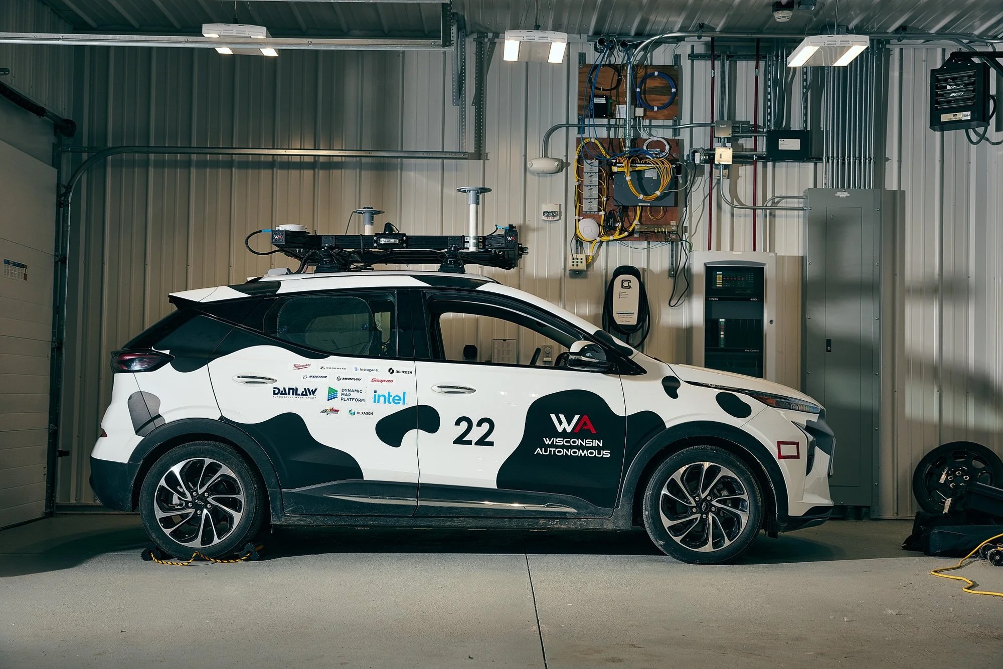 The Wisconsin Autonomous team's black-and-white cow-patterned self-driving Chevrolet Bolt, number 22, with a roof-mounted sensor array, parked in a garage.